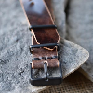 May include: Close-up of a brown leather watch strap with black hardware. The strap has a rich, textured appearance with visible stitching and a matte black buckle. The leather shows variations in color.