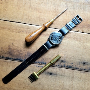 May include: A black leather watch strap with stitching, a wood handled awl, and a brass leather punch tool on a wooden surface.