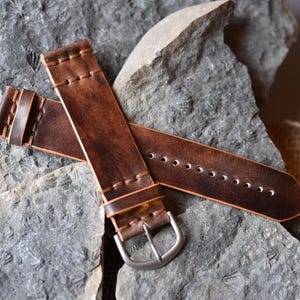 May include: Two brown leather watch straps with a distressed finish. The straps are stitched with a contrasting thread and feature a silver-colored buckle. The straps are displayed on a gray stone surface.