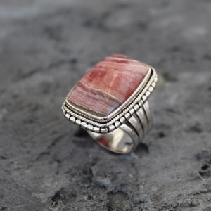 Könnte beinhalten: Ein silberner Ring mit einem rechteckigen rosa und weißen Edelstein, der in einer Perlenfassung sitzt. Der Ring hat ein breites Band mit mehreren Rillen.