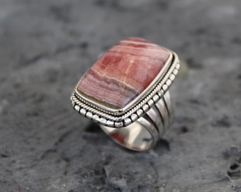 Pode incluir: Um anel de prata com uma pedra rectangular rosa e branca engastada em uma moldura de contas. O anel tem uma banda larga com múltiplos sulcos.