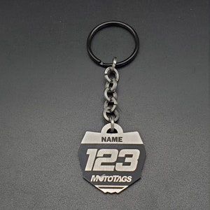 May include: A black and silver metal keychain with a number plate design. The plate has the number "123" and the text "NAME" and "MOTOTAGS".