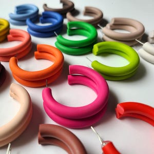 May include: A collection of colorful hoop earrings in various shades, including pink, orange, green, and blue. The earrings are made of a smooth, solid material and are arranged on a white surface. Each earring has a post for pierced ears.