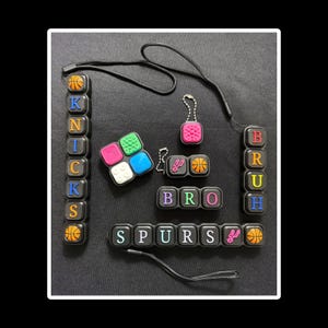 May include: A collection of black silicone sensory fidget accessories featuring colorful letters spelling KNICKS, BRUH, BRO, and SPURS, along with basketball icons, a San Antonio Spurs logo, and various textured square shapes, some attached to black lanyards.