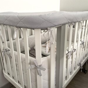 May include: A white crib with a gray quilted bumper pad. The crib has a white wooden frame and a white mattress. The bumper pad is tied to the crib with gray ribbons. The crib is in a room with white walls.