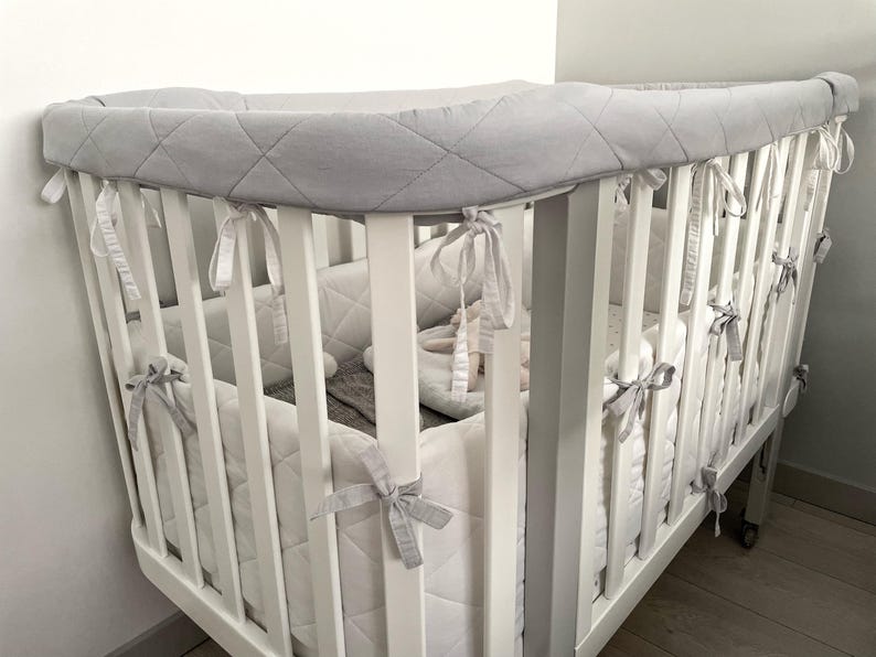 May include: A white crib with a gray quilted bumper pad. The crib has a white wooden frame and a white mattress. The bumper pad is tied to the crib with gray ribbons. The crib is in a room with white walls.