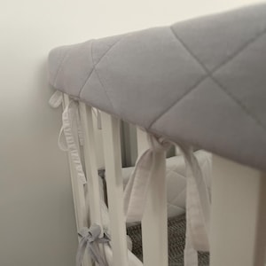 May include: A white crib with a gray quilted bumper pad. The bumper pad has white ties that are tied around the crib rails.