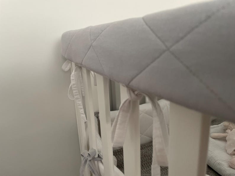 May include: A white crib with a gray quilted bumper pad. The bumper pad has white ties that are tied around the crib rails.