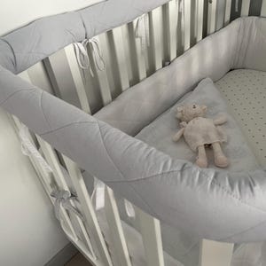 May include: A white crib with a gray quilted bumper pad and a white sheet with gray polka dots. A pink stuffed animal is lying on the sheet.