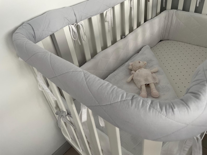 May include: A white crib with a gray quilted bumper pad and a white sheet with gray polka dots. A pink stuffed animal is lying on the sheet.