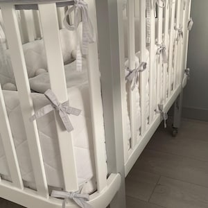 May include: A white crib with a gray quilted bumper pad. The crib has a white wooden frame and a gray fabric bumper pad with ties. The crib is on a wooden floor.