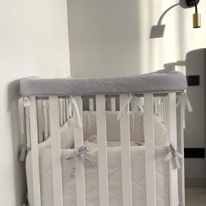 May include: White oval crib with a gray quilted bumper pad. The crib has white slats and a white mattress. The crib is on wheels and has a gray quilted bumper pad with white ties.