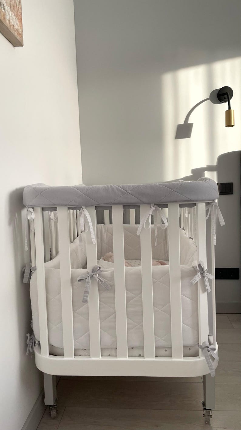 May include: White oval crib with a gray quilted bumper pad. The crib has white slats and a white mattress. The crib is on wheels and has a gray quilted bumper pad with white ties.