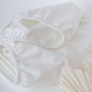 Peut inclure: Une couche en tissu réutilisable en coton blanc avec des élastiques autour des jambes et de la taille. La couche est pliée et roulée.