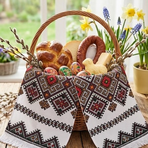 Könnte beinhalten: Ein geflochtener Korb gefüllt mit Osterleckereien, darunter ein geflochtener Brotlaib, Käse, Wurst und verzierte Eier. Der Korb ist mit einem weißen, bestickten Tuch mit geometrischem Muster ausgekleidet. Narzissen und Weidenkätzchenzweige sorgen für Frühlingsstimmung.