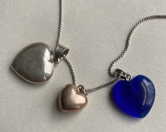 Puede incluir: Un collar de cadena de plata con tres colgantes en forma de corazón. Un colgante es de plata, uno es de oro y uno es de vidrio azul.