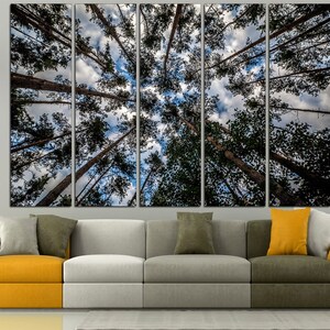 May include: A multi-panel canvas print depicting a view looking up through a forest canopy. Tall, slender tree trunks reach towards a cloudy sky with patches of blue. The artwork is displayed above a modern sectional sofa.