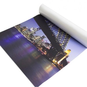 Puede incluir: Impresi&oacute;n enrollada de un paisaje urbano nocturno, con un puente y edificios iluminados. La impresi&oacute;n tiene una superficie blanca y brillante en el exterior y un interior azul. La imagen muestra una escena nocturna vibrante con luces de la ciudad reflejadas en el agua.