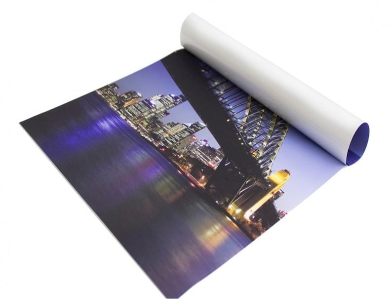 Puede incluir: Impresi&oacute;n enrollada de un paisaje urbano nocturno, con un puente y edificios iluminados. La impresi&oacute;n tiene una superficie blanca y brillante en el exterior y un interior azul. La imagen muestra una escena nocturna vibrante con luces de la ciudad reflejadas en el agua.