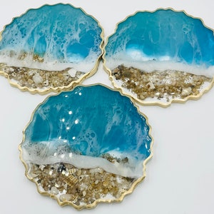 May include: Three round resin coasters featuring a beach scene. The coasters depict a blue ocean with white foam waves, a white sandy beach, and brown and white seashells. The coasters have a gold rim.