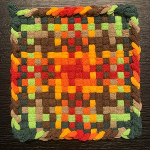 May include: A woven square potholder with a colorful geometric pattern in shades of green, brown, orange, red, and yellow. The potholder has a braided border.