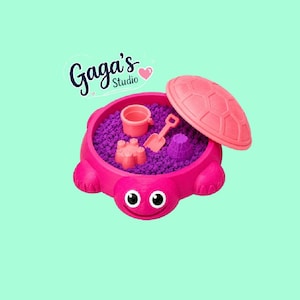 May include: A bright pink turtle-shaped sandbox filled with purple kinetic sand, featuring a matching turtle shell lid, a small pink bucket, a pink shovel, and two sand molds. The text Gaga's Studio with a small pink heart is visible above.