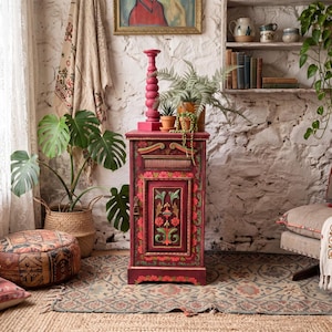 May include: A tall, rectangular wooden cabinet painted in deep red with intricate hand-painted floral motifs in green, yellow, and orange. The cabinet features a single door with a central carved panel, a small drawer at the top, and a metal latch.