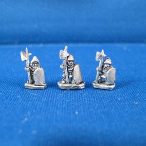 Puede incluir: Tres figuras metálicas en miniatura de soldados medievales, cada una con una alabarda y un casco y escudo. Las figuras están pintadas en plata.