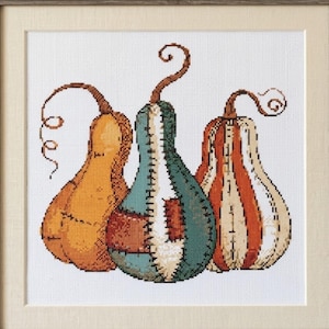 May include: A framed cross-stitch artwork featuring three stylized gourds. The left gourd is orange, the center is teal and white with patchwork details, and the right is orange and cream striped. Each gourd has a brown stem with a curly vine.