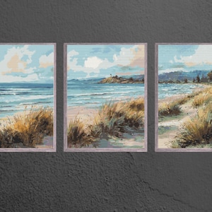 May include: Triptych of framed beach landscape paintings. Each painting shows a coastal scene with a blue ocean, sandy beach, and grassy dunes under a cloudy sky. The artwork is framed in a light-coloured wood.