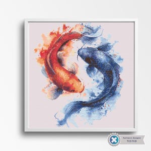 May include: A framed cross-stitch artwork featuring two koi fish in a yin-yang composition. One fish is red and orange, the other is blue. The background is a light pink, and the frame is white.