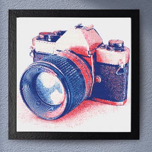 Op de afbeelding: Ingelijste kunstprint van een vintage camera in pop-artstijl. De camera is weergegeven in tinten rood en blauw, met een zwart frame. De afbeelding is een close-up van de camera, waarbij de lens en de body worden benadrukt.