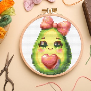 May include: A cross-stitch embroidery hoop with a cute avocado design. The avocado is green with a pink bow and a pink heart. The embroidery is on white fabric.