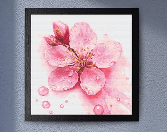 Puede incluir: Una impresión enmarcada en acuarela de una flor de cerezo rosa con gotas de agua. La flor tiene múltiples pétalos y un centro amarillo. La obra de arte está en un marco negro y se muestra contra una pared azul claro.