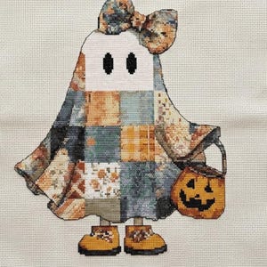 This Halloween cross stitch pattern show a sweet ghost character crafted with detailed patchwork textures and warm fall colors. The ghost is decorated with quilt-inspired blocks in shades of orange, soft blue, ivory, and charcoal grey, accented by a large coordinating bow. A smiling pumpkin candy bucket completes the festive look. Perfect for stitchers seeking a cute ghost embroidery pattern, cottagecore Halloween decor, autumn craft project, or charming seasonal art with a cozy handmade feel.
