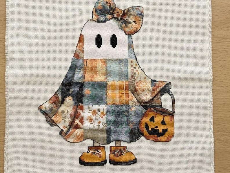 This Halloween cross stitch pattern show a sweet ghost character crafted with detailed patchwork textures and warm fall colors. The ghost is decorated with quilt-inspired blocks in shades of orange, soft blue, ivory, and charcoal grey, accented by a large coordinating bow. A smiling pumpkin candy bucket completes the festive look. Perfect for stitchers seeking a cute ghost embroidery pattern, cottagecore Halloween decor, autumn craft project, or charming seasonal art with a cozy handmade feel.