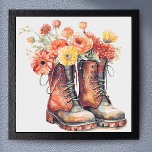 May include: A framed print featuring a pair of brown work boots filled with colorful flowers. The boots have laces and eyelets, and the flowers include orange, yellow, and red blooms. The artwork is set against a white background.