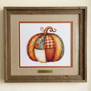 Könnte beinhalten: Ein gerahmtes Kreuzstichbild, das einen Kürbis mit einem Patchwork-Design aus orangefarbenen, braunen und blauen Stoffmustern zeigt. Das Werk befindet sich in einem rustikalen Holzrahmen mit beigem Passepartout und einem Schild mit der Aufschrift The Patchwork Harvest.