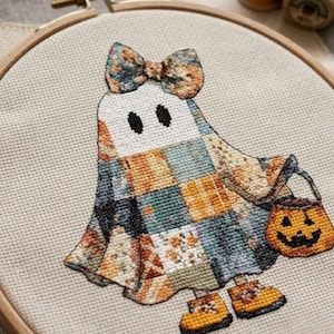 Puede incluir: Un proyecto de bordado en punto de cruz en un bastidor de madera que muestra un fantasma con un patrón de patchwork en tonos azules, naranjas y blancos. El fantasma lleva un lazo a juego, sostiene un cubo de calabaza y calza botas amarillas.