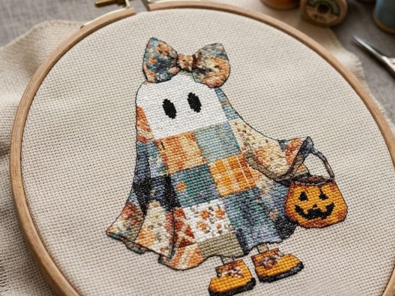A Halloween ghost cross stitch pattern of a friendly ghost dressed in a beautiful patchwork costume made from soft autumn fabrics in muted orange, cream, blue, grey, and golden tones. The ghost wears a large matching bow and tiny yellow shoes while carrying a cheerful jack-o'-lantern treat bucket. This cottagecore Halloween design creates a warm and inviting seasonal atmosphere, making it ideal for autumn decor, Halloween home decoration, handmade gifts, and relaxing seasonal stitching projects.