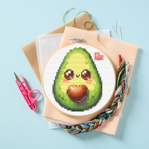 May include: A cross-stitch embroidery hoop with a cute cartoon avocado with a heart in its hand. The avocado is green with a brown pit and has a smiling face with big eyes. The background is white with a pink heart in the upper right corner.