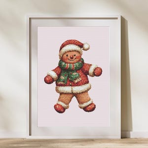 May include: Framed print of a watercolour gingerbread man illustration. The gingerbread man is dressed in a red Santa hat, coat, and mittens, with a green and red scarf. The artwork has a soft, textured look and is set against a pale pink background.