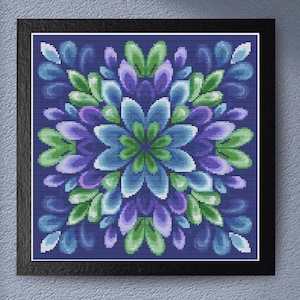 Puede incluir: Una obra de arte de punto de cruz enmarcada con un diseño floral simétrico. El diseño presenta una flor central en tonos verdes y azules, rodeada de pétalos en tonos azules, morados y verdes, sobre un fondo azul oscuro.