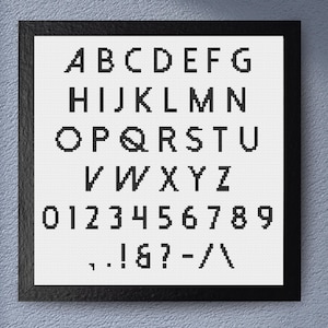 Op de afbeelding: Een ingelijste kruissteek sampler met het alfabet, cijfers en leestekens. De zwarte borduursels staan op een witte achtergrond, met een zwarte lijst. Het lettertype is eenvoudig en modern.