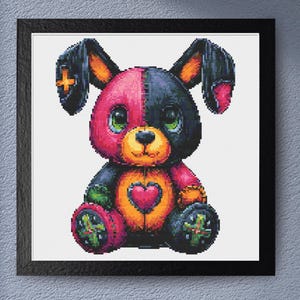 May include: A framed cross-stitch pattern featuring a split-colour plush toy with one side in bright pink and the other in dark black. The toy has large green eyes, a yellow muzzle, a central pink heart on its chest, and green accents on its feet.