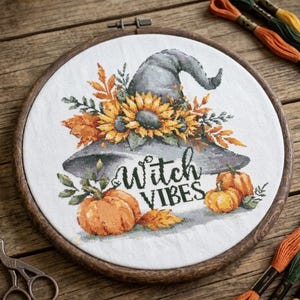 May include: A circular wooden embroidery hoop containing white fabric with a cross-stitch design of a grey witch hat adorned with yellow sunflowers and orange autumn leaves, featuring two orange pumpkins and the text Witch VIBES in black.