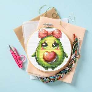 May include: A cross-stitch embroidery hoop with a green avocado design. The avocado has a pink bow and a pink heart in its mouth. The avocado has a cute cartoon face with big eyes.