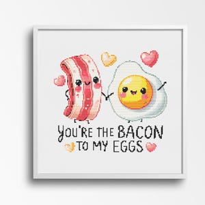 May include: A framed cross-stitch featuring a cartoon bacon strip and a fried egg, both with smiling faces, holding hands. The text "You're the bacon to my eggs" is below them, with heart and star accents.