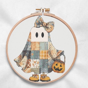 A charming Halloween ghost cross stitch of a friendly ghost dressed in a  patchwork costume of warm autumn colors in soft oranges, creamy neutrals, muted blues, and rustic greys. Accented with a large coordinating bow, tiny yellow shoes, and a cheerful jack-o'-lantern treat bucket, this cottagecore Halloween design captures the warmth and whimsy of the season. Perfect for autumn decorating, seasonal decor, handmade gifts, and relaxing cross stitch projects that bring festive charm to any home.
