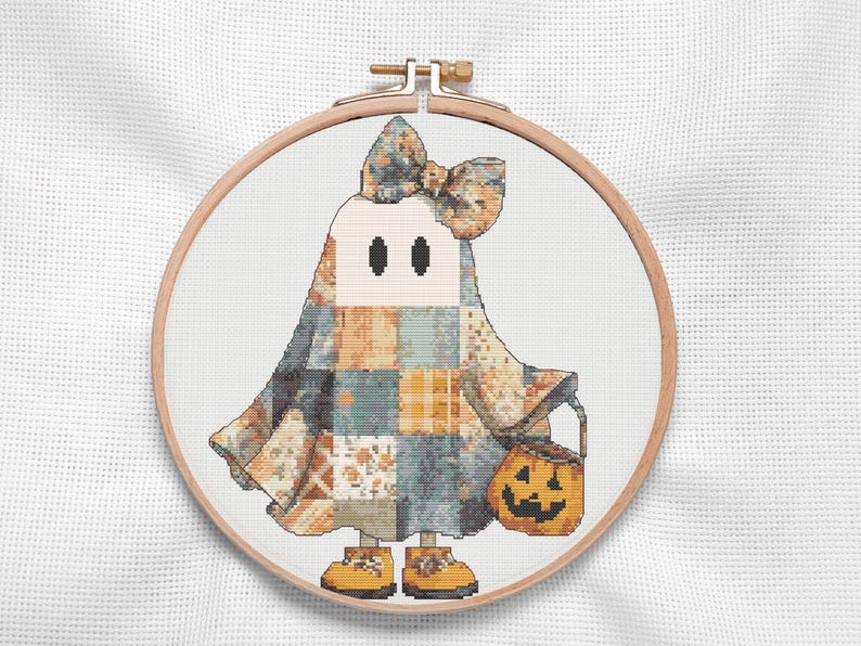 A charming Halloween ghost cross stitch of a friendly ghost dressed in a  patchwork costume of warm autumn colors in soft oranges, creamy neutrals, muted blues, and rustic greys. Accented with a large coordinating bow, tiny yellow shoes, and a cheerful jack-o'-lantern treat bucket, this cottagecore Halloween design captures the warmth and whimsy of the season. Perfect for autumn decorating, seasonal decor, handmade gifts, and relaxing cross stitch projects that bring festive charm to any home.